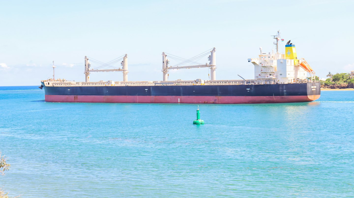 Cargo ship sails past a green navigational buoy near a coastline.