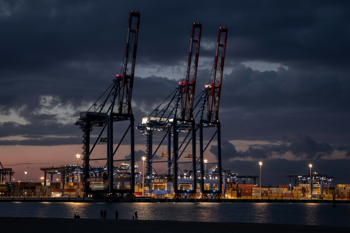 Container cranes and stacked shipping containers at a busy port at dusk.